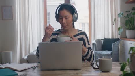 アジアの若い女性がラップトップコンピュータを使用し,自宅で学生のブレインストーミングプロジェクトを実行し,オンラインで情報を調査し,朝食を食べ,勉強を楽しんでいます