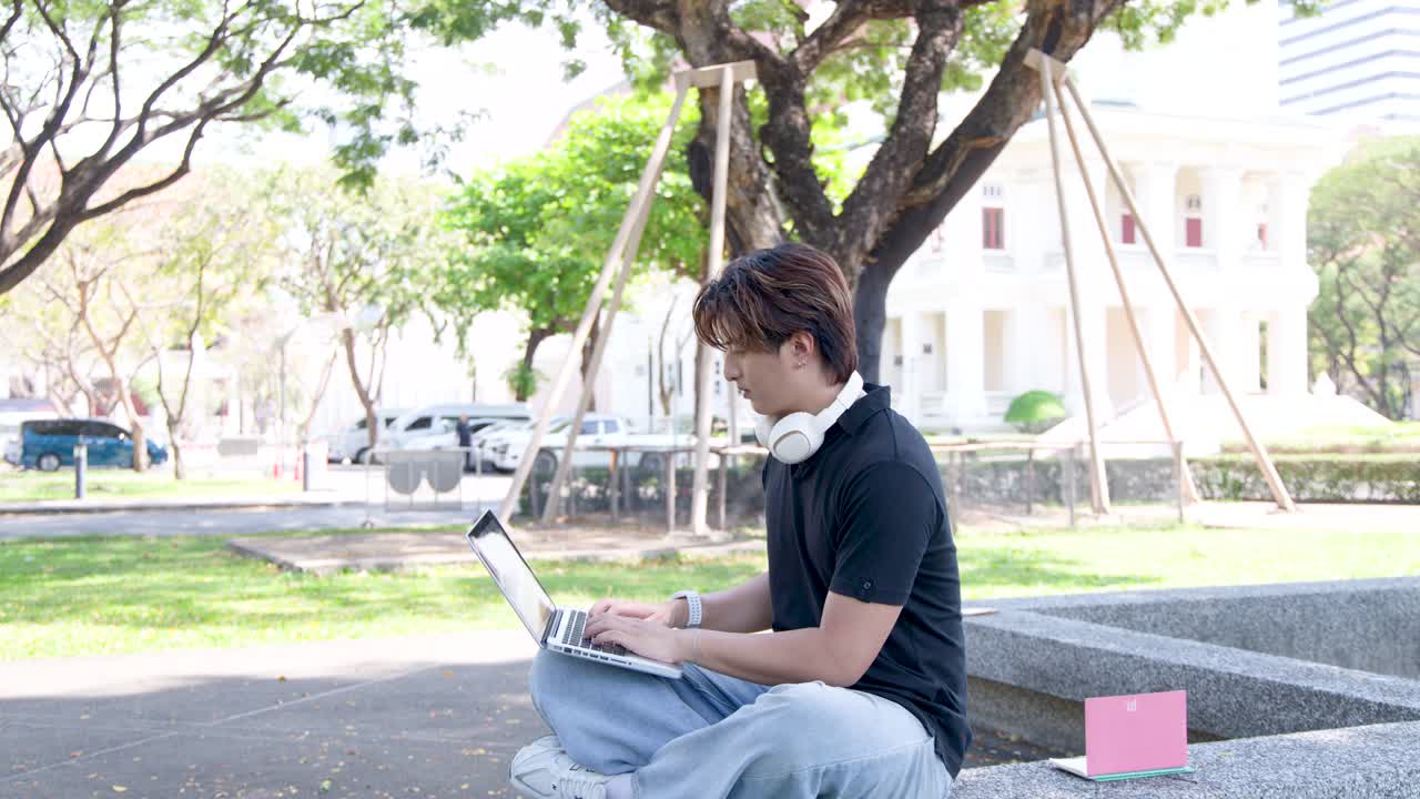 A young man sits outdoors on a campus ledge using a laptop under bright sunlight