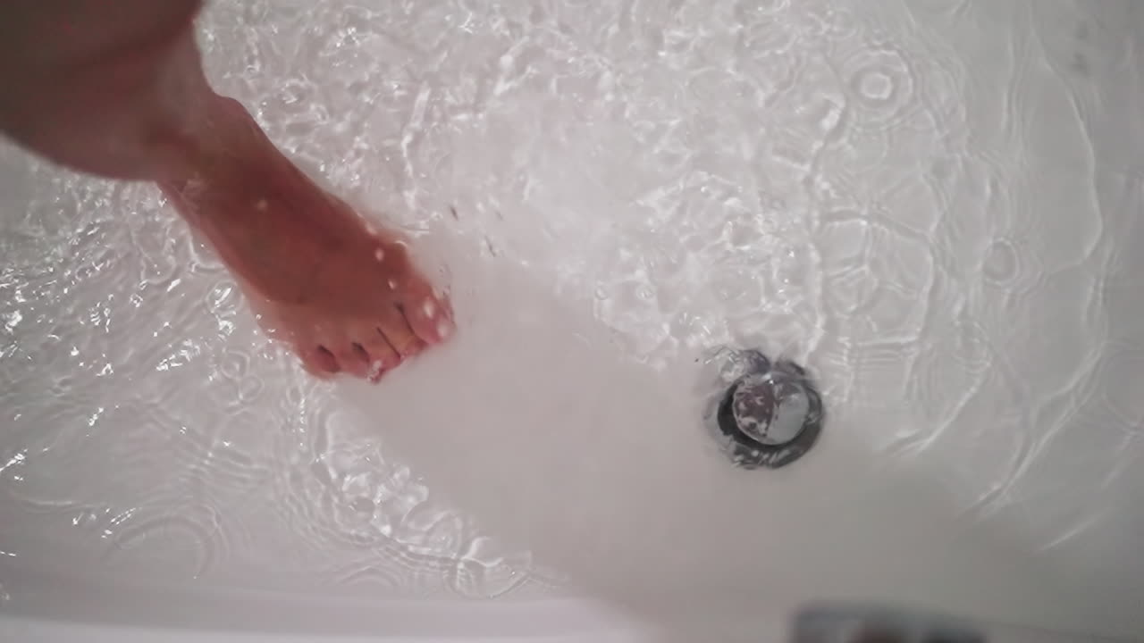 Woman stands in bathtub with dripping water closeup. Lady foot in clear liquid and falling shower drops in bathroom. Woman enjoys cozy bath