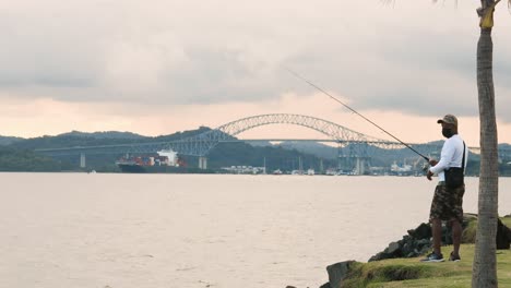 Mann-Wirft-Stark-Angelhaken-Ins-Wasser-Und-Versucht,-Einen-Zu-Fangen,-Um-Mit-Seiner-Familie-Zu-Essen,-Während-Er-Die-Zeit-Allein-Mit-Blick-Auf-Die-Brücke-Der-Amerika-Und-Die-Schiffe-Genießt,-Die-Den-Panamakanal-Durchqueren