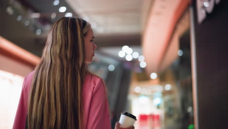 back view of woman in pink dress holding coffee cup while walking through brightly lit mall, the blurred background features stores and mall lights with bokeh effect and unclear items