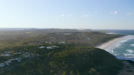 aerial shot of forest-covered coastline, with residential buildings nestled along the shore. waves crash onto the rocks, capturing the natural beauty of the landscape at sunset