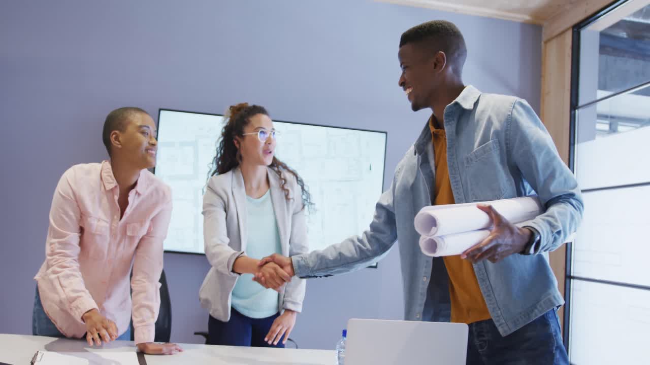 Three diverse male and female architects standing at table shaking hands in meeting room