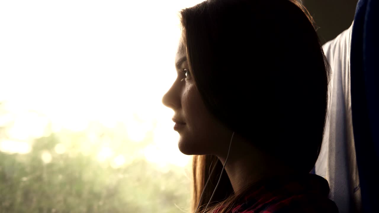 chica atractiva sentada en la sombra en el tren. escuchando música con auriculares y teléfono móvil. rayos de sol en la ventana. paisaje natural afuera
