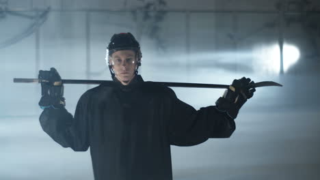 retrato de un joven jugador de hockey sosteniendo un club sobre los hombros y mirando la cámara en la pista de hielo