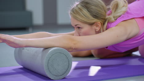 lady in pink outfit engages in a flexibility workout using a foam roller with the assistance of another lady in brown, fitness ball visible in the gym background