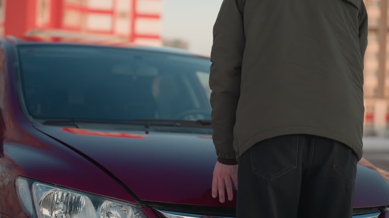 Back view of boy in green winter jacket closing car bonnet during cold day, two people standing and interacting in blurred background, snowy urban setting with red building and trees visible