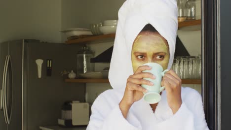 Fröhliche-Afroamerikanische-Frau-Mit-Schönheitsmaske-Im-Gesicht,-Die-In-Der-Küche-Kaffee-Trinkt