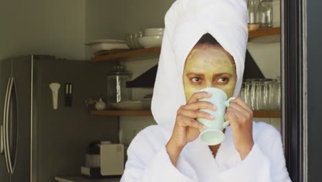 gelukkige afro-amerikaanse vrouw met schoonheidsmasker op het gezicht, koffie drinken in de keuken