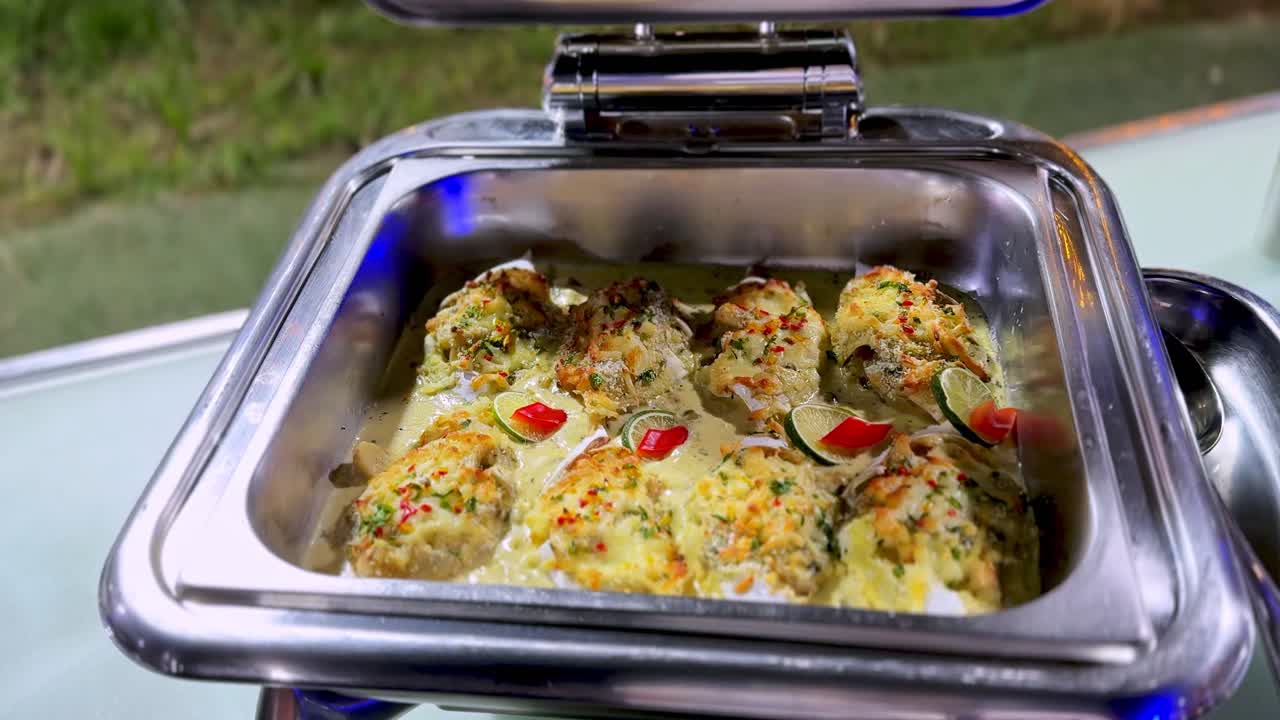 Stainless steel warmer holding baked fillets topped with melted cheese and herbs. Creamy sauce base with lime and red pepper garnish at an outdoor garden buffet or wedding reception