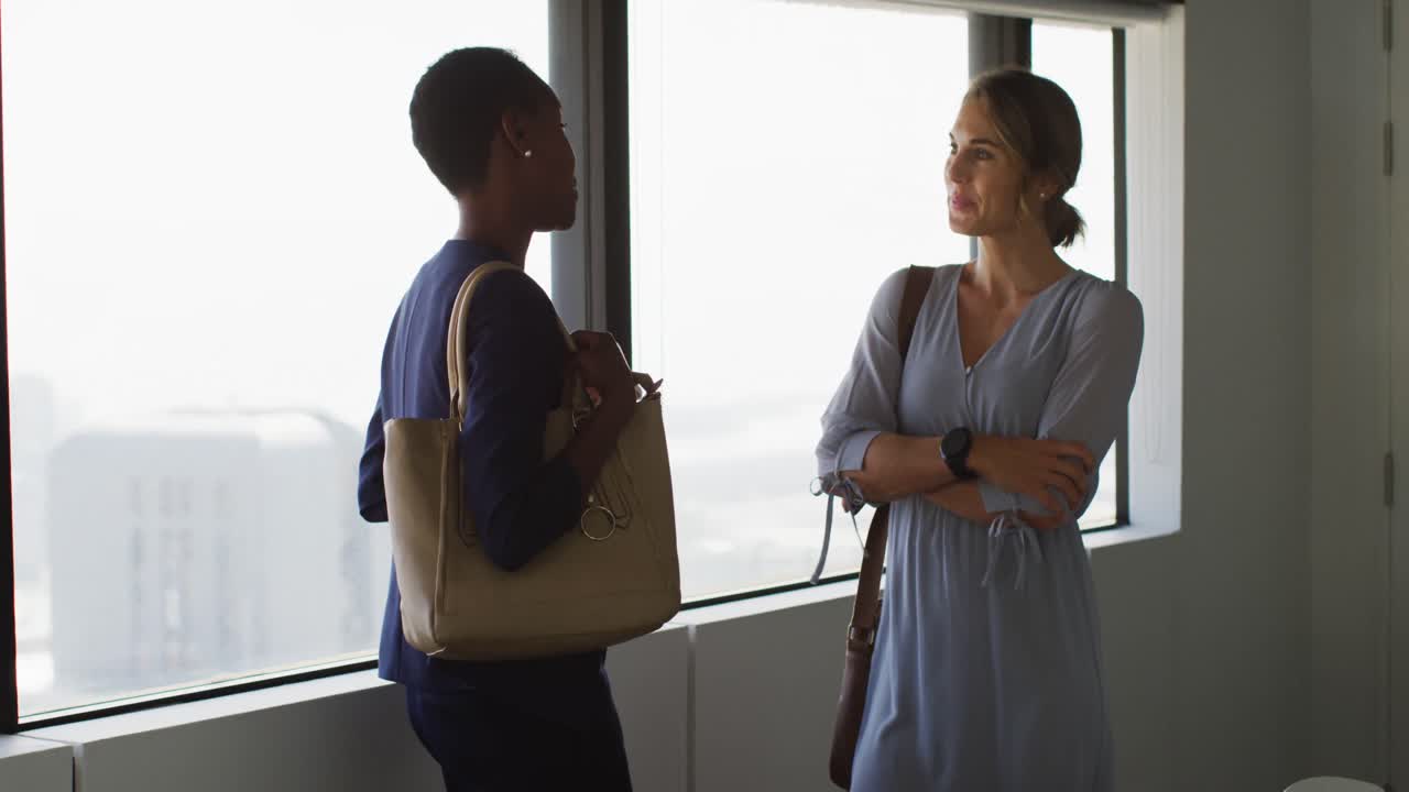 두 명의 다양한 여성 동료가 창문에 서서 이야기하고 미소 짓고 있습니다.