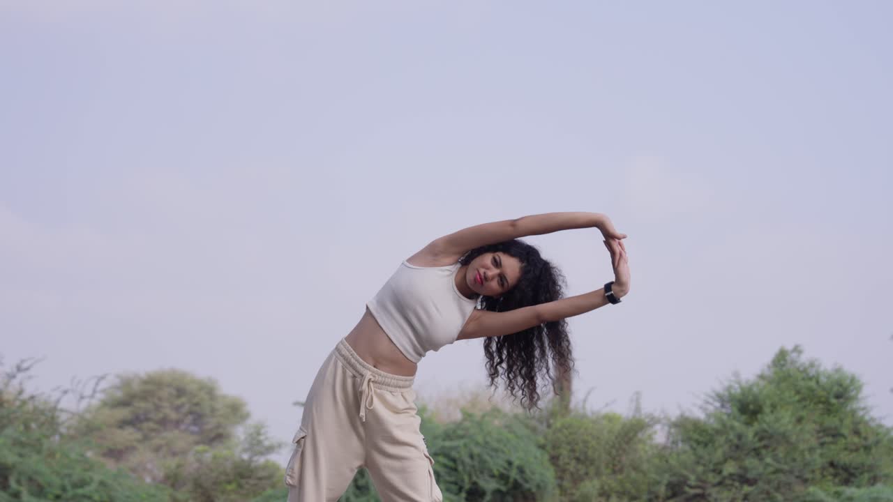 A slow-motion of a South Asian young woman with curly hair training outdoors, with trees and sky behind her. Taken from a low angle