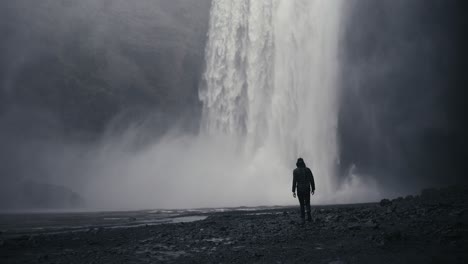 Wide-shot-of-a-dark-lone-hiker-walks-towards-an-epic-downpour-from-a-water-fall-amidst-dark-and-moody-rocks