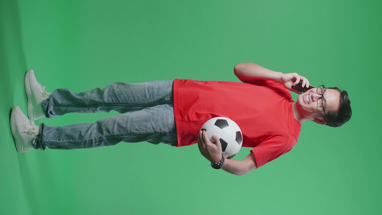 녹색 화면 배경에서 축구를 응원하는 동안 스마트폰으로 말하는 공을 가진 아시아 남자의 몸이 가득 차 있습니다.