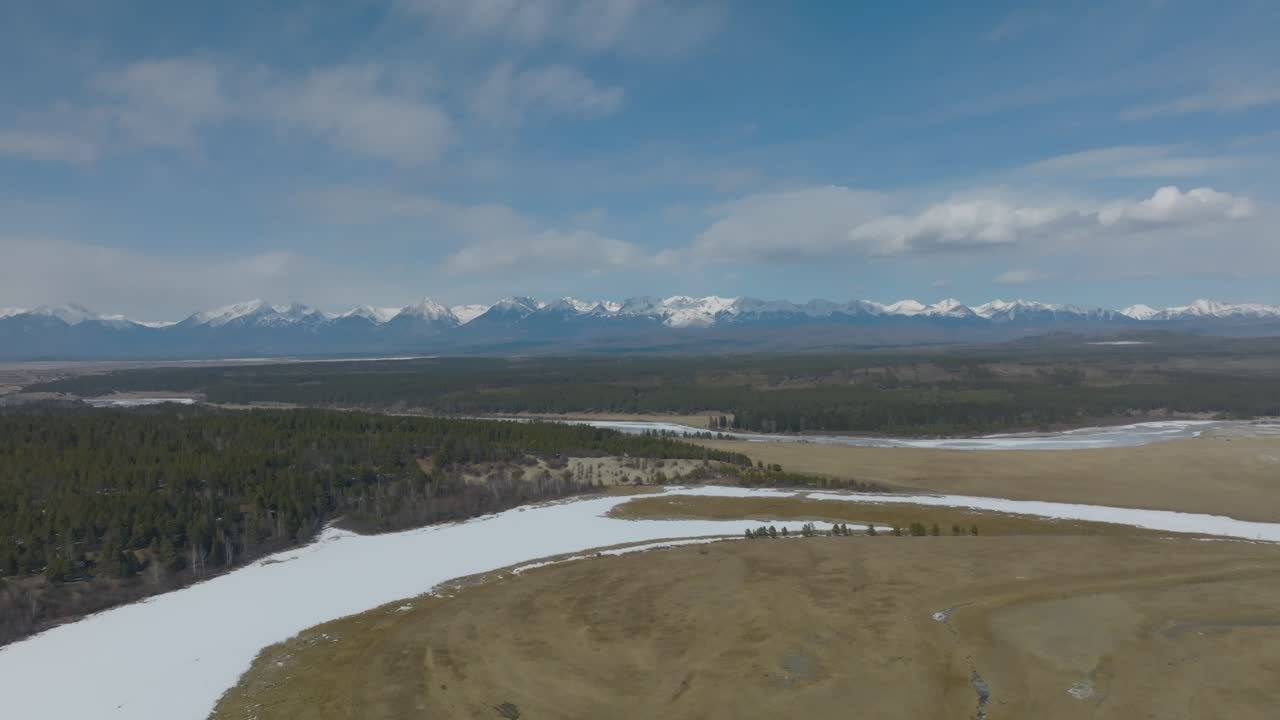 Rekaman drone sungai beku yang lebar berkelok-kelok melalui hutan dan stepa dengan pegunungan bersalju di kejauhan di bawah langit biru di Buryatia, Siberia, Rusia
