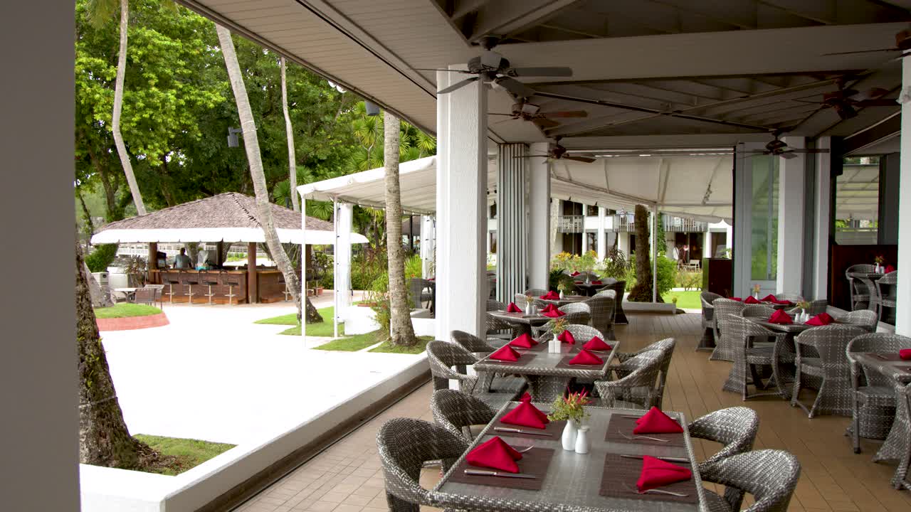Plano general fijo de un restaurante vacío de un resort tropical con servilletas rojas y sillas de mimbre.