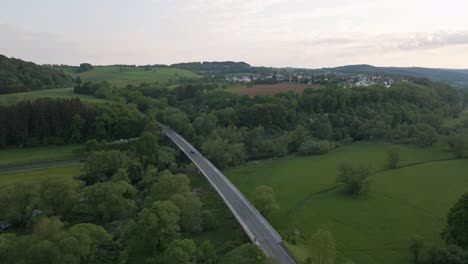 Static-aerial-view-of-a-dark-car-that-crosses-a-bridge-that-leads-into-a-lush-deciduous-forest