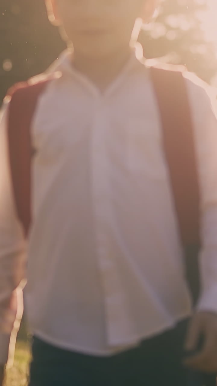 joyful junior schoolboys in shirts with backpacks walk after lessons in green park at back sunlight slow motion