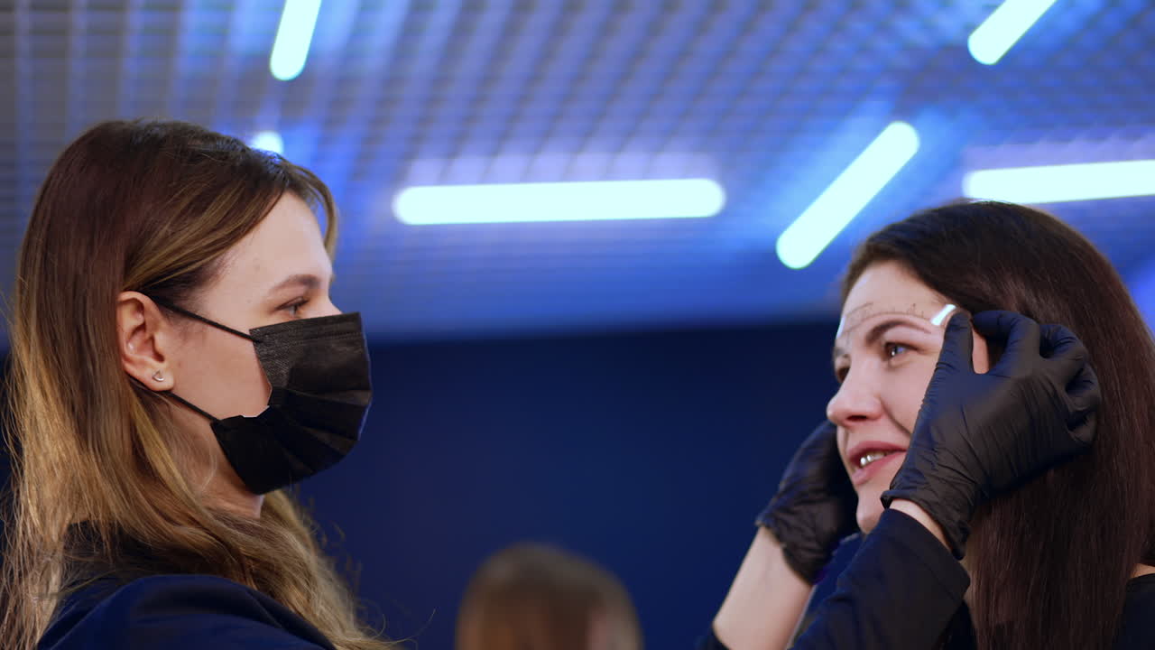 Female cosmetologist stuck the ruler to a brunette woman's forehead. The client is prepared for permanent eyebrow make up.