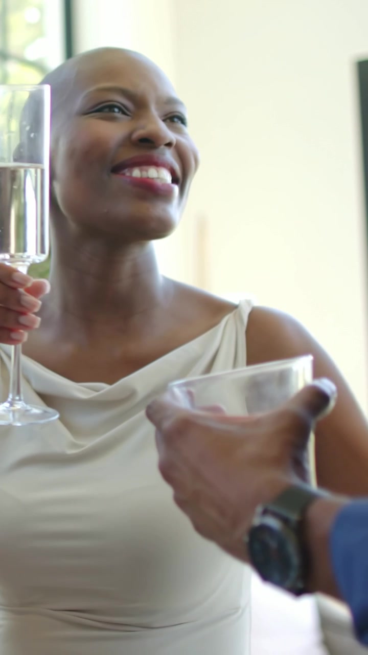 Vertical video: Reaching man with drink, woman in cream dress clinking flute by window, celebrating