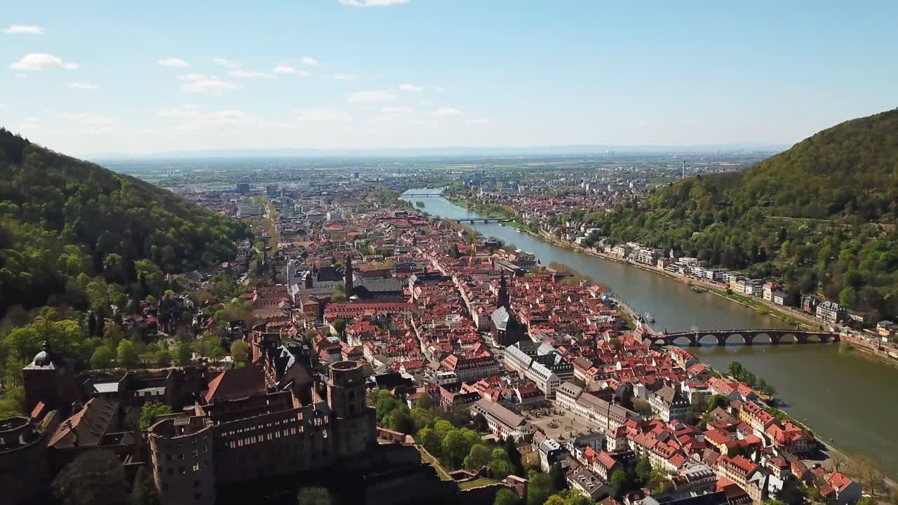 A stunning aerial view of Heidelberg, Germany, showcasing the historic Old Town, Neckar River, red-roofed buildings, and lush green hills. The Heidelberg Castle is visible in the foreground