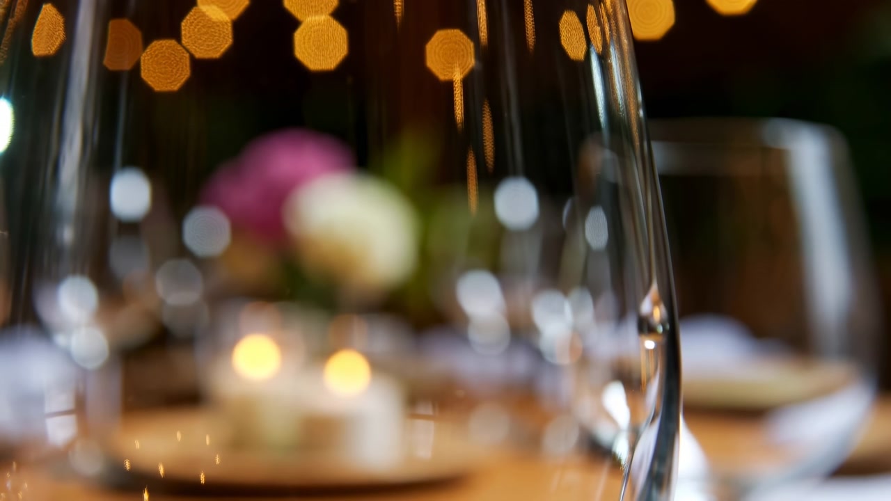 Un calice da vino in primo piano con una piccola goccia che scivola lungo la superficie interna lascia una scia debole, mentre luci calde sullo sfondo creano un bokeh morbido, riflessi lucidi, rifrazione delicata e una cena romantica.