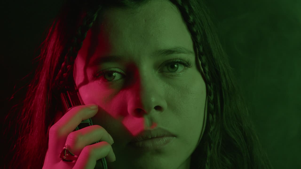 A close-up shot of a woman holding a phone, with striking red and green lighting creating a mysterious atmosphere. Her expression is thoughtful and intense, enhancing the mood