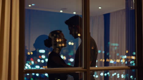romantic couple silhouetted against city night view from apartment window