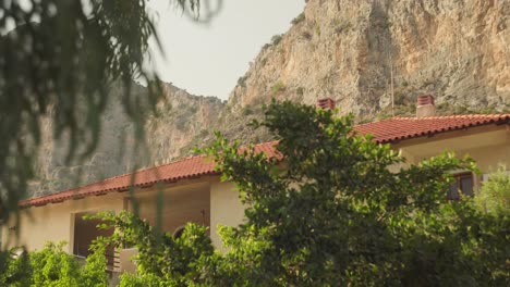 wind blows green tree and shrub as sunlight illuminates house below towering cliffs of leonidio greece