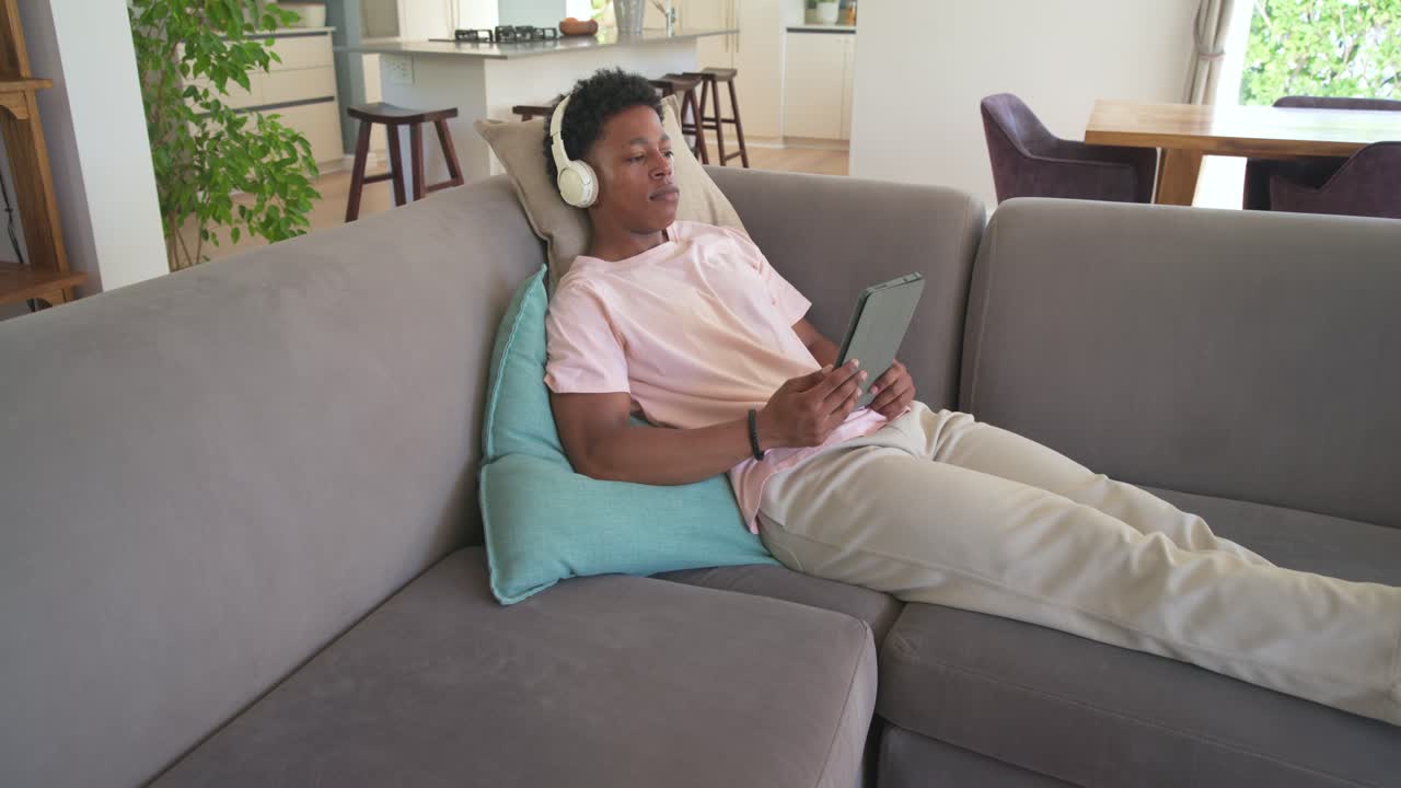 Uomo afroamericano sdraiato su un divano grigio con cuffie bianche, che tocca un tablet e sorride allo schermo.
