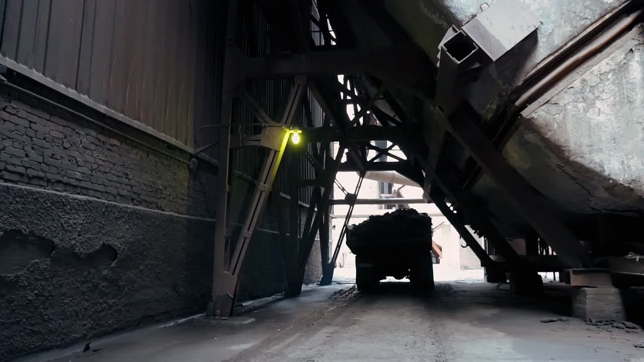 Large dump truck hauls construction debris down a dirt road from an old abandoned factory. Rusty destroyed structures and walls of the power plant