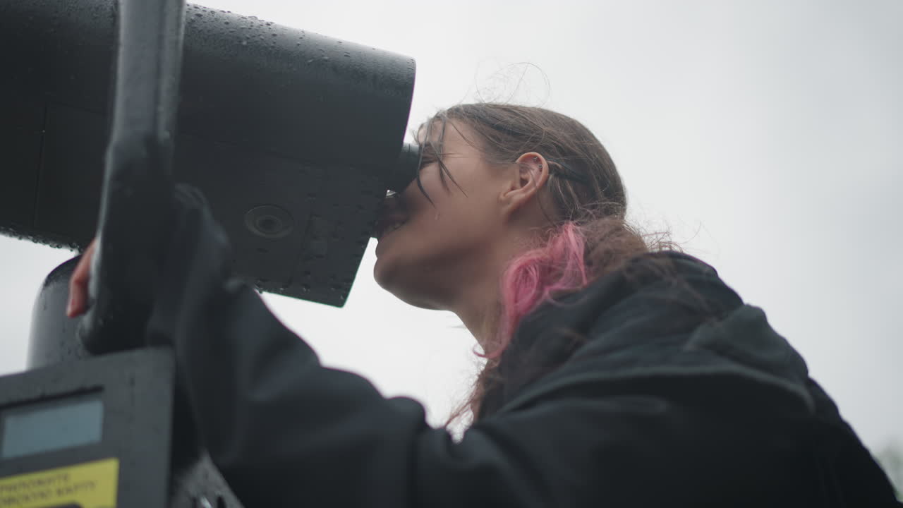 Giovane donna che guarda attraverso un visore su un lungomare nuvoloso, gocce di pioggia sui capelli e sulla giacca, primo piano dal basso, studentessa contemplativa che esplora lo skyline lontano con un binocolo bagnato, cinematografico.