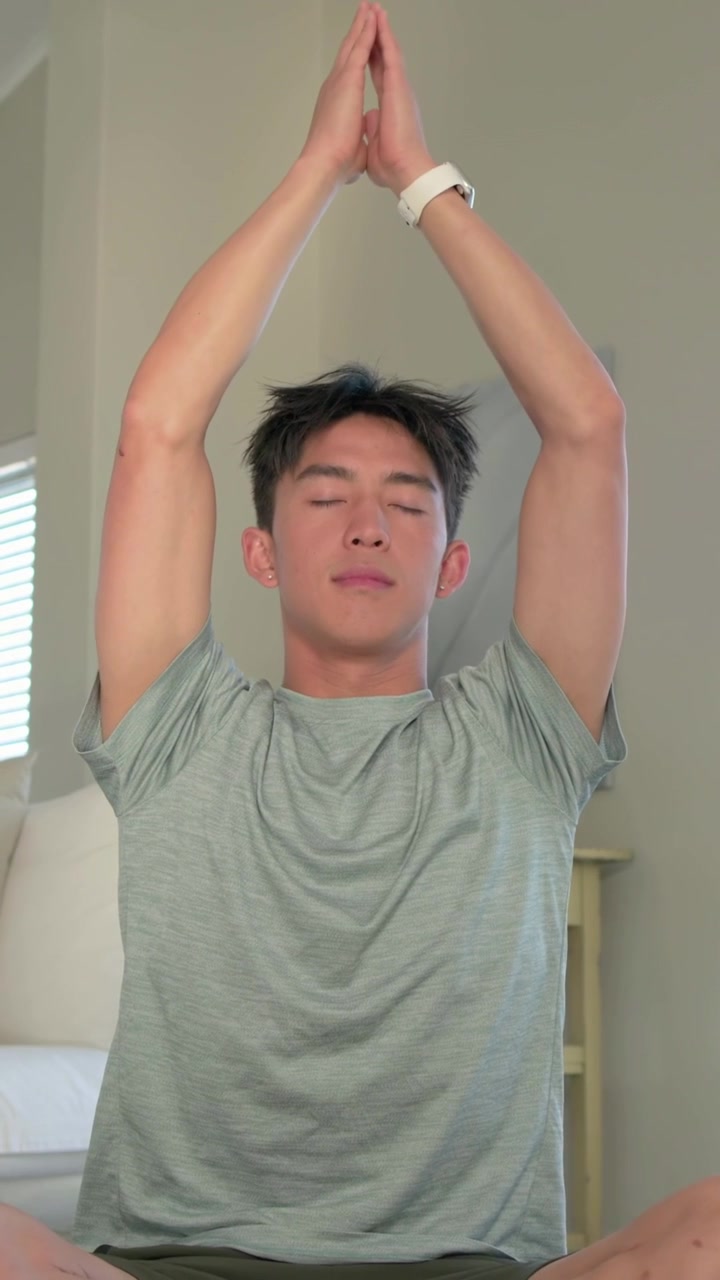 縦型動画：セッションを開始するグレーのTシャツを着た男性が、ソファで腕を上げ、合掌して祈っている。時計。