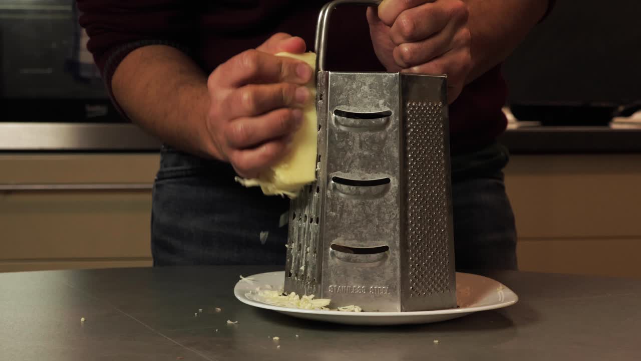 A close-up of hands grating fresh cheese using a stainless steel grater in a kitchen setting. Perfect for illustrating food preparation, cooking, and the use of fresh ingredients in recipes.