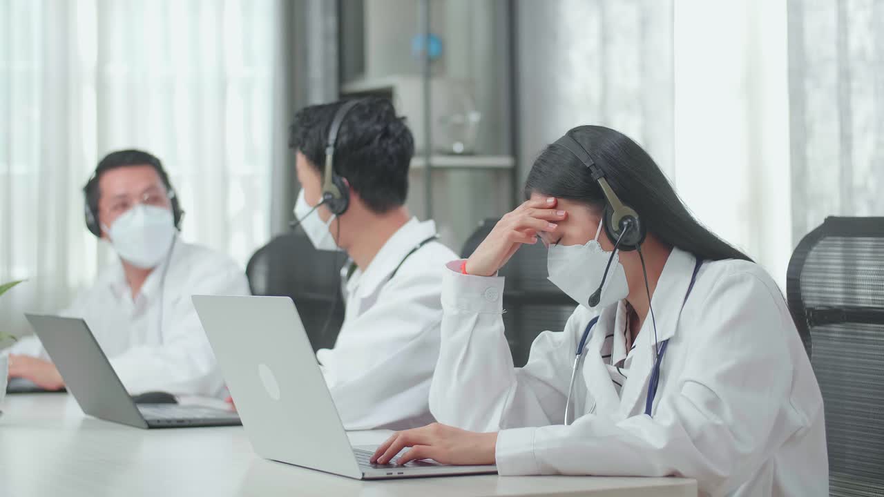 A Woman Of Three Asian Doctors With Stethoscopes In Headsets And Masks Working As Call Center Agent Is Tired Because Her Colleagues Are Talking During Working At The Office