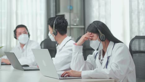 a woman of three asian doctors with stethoscopes in headsets and masks working as call center agent is tired because her colleagues are talking during working at the office