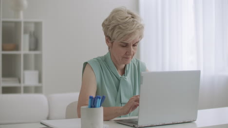 aged-blonde-woman-is-browsing-internet-and-typing-message-at-keyboard-of-laptop-sitting-at-her-home-at-weekend