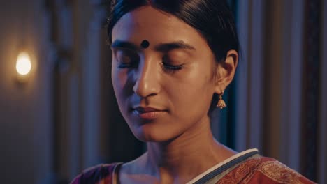 in the serene ambiance of a dimly lit temple, a young indian woman, adorned in traditional attire, closes her eyes, immersing herself in a moment of quiet contemplation and spiritual connection