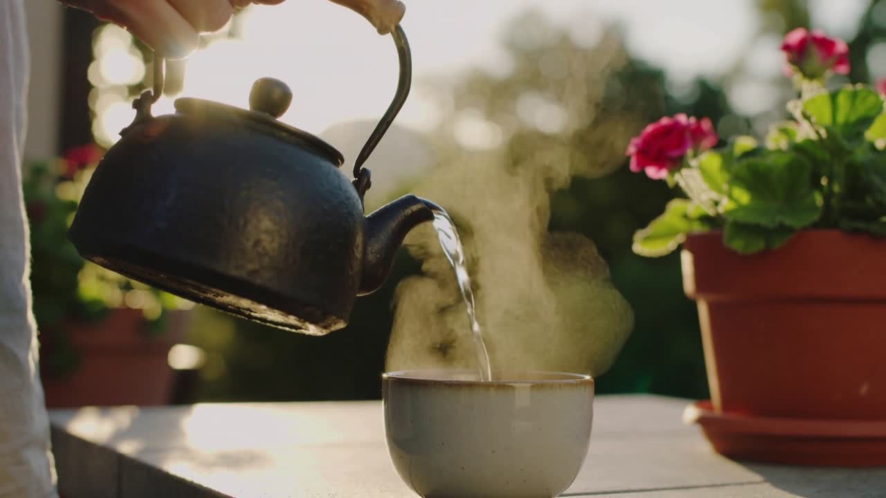 Una mano adulta inclina un bollitore di metallo, riempiendo una tazza di ceramica su un patio, con vapore che sale e fiori in vaso.