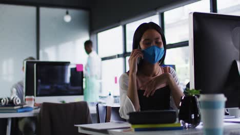 mujer asiática con máscara facial hablando en un teléfono inteligente mientras está sentada en su escritorio en una oficina moderna
