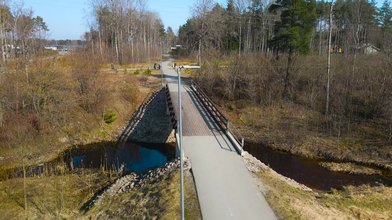 Aerial pan around wooden footbridge over slope river bank, bridge connects with a asphalt pedestrian path in Laagri. Long empty walkway stretches across nature park walking trail with trees all around