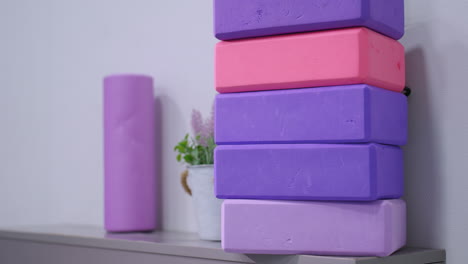 stacked colorful yoga blocks in shades of purple and pink neatly arranged on cabinet with a matching foam roller and potted plant in background, creating a calming and organized fitness studio setting