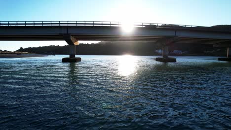 drone flying low under road bridge at sunset over glittering water