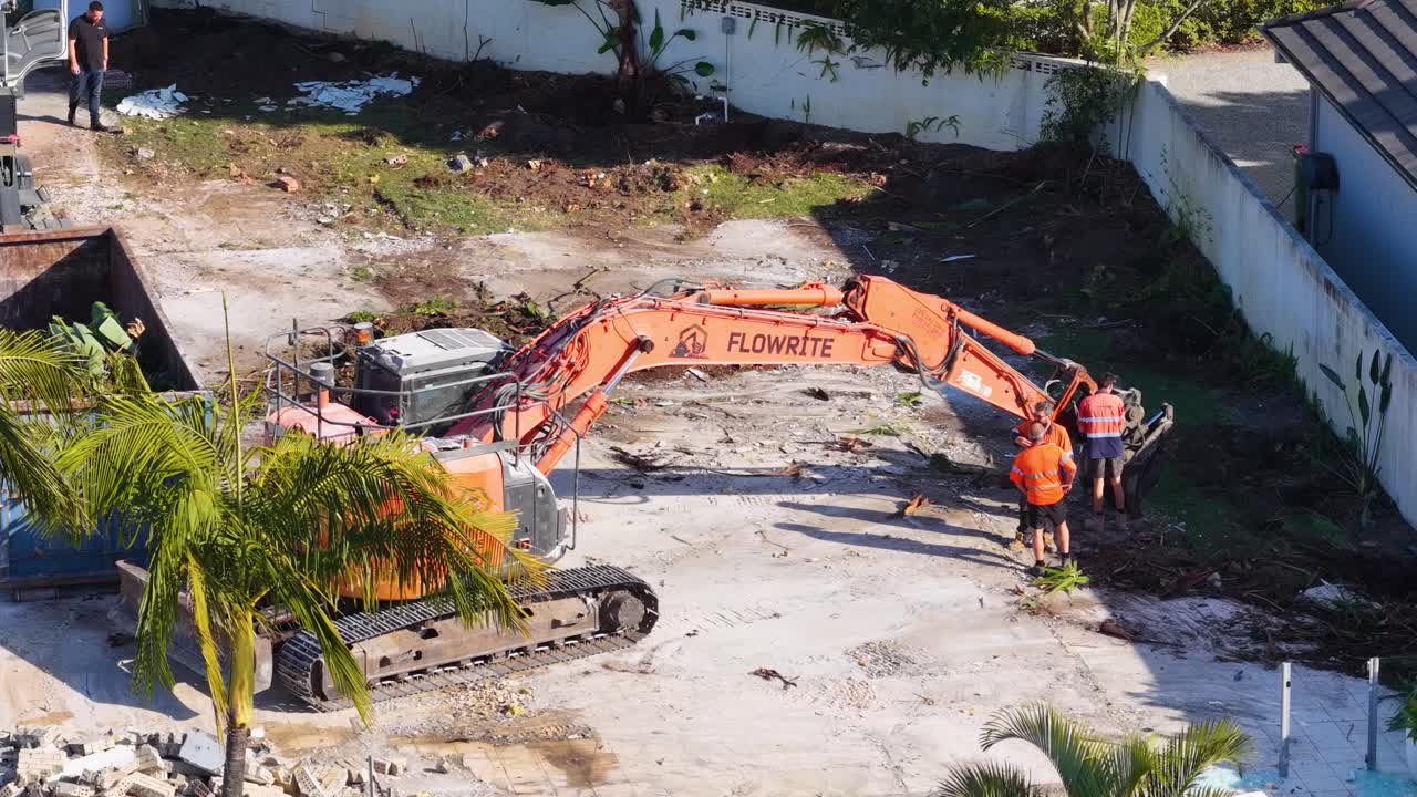 Two workers in high-visibility clothing direct an orange excavator on a sunny demolition site, clearing debris from a former residential property in Gold Coast