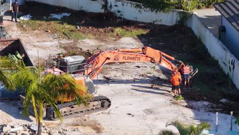 two workers in high-visibility clothing direct an orange excavator on a sunny demolition site, clearing debris from a former residential property in gold coast