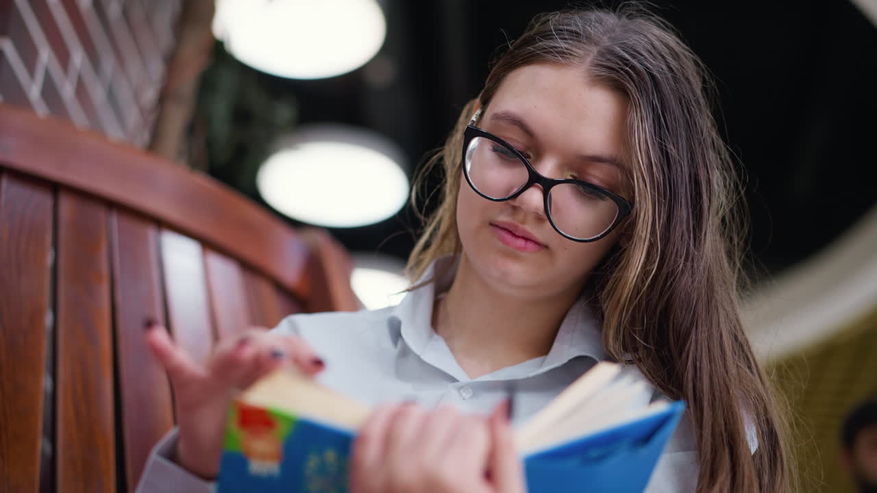 la dama con gafas abre cuidadosamente una nueva página, la cabeza inclinada hacia un lado, centrada en la lectura, tres grandes luces brillan en el fondo borroso, creando un ambiente cálido y moderno