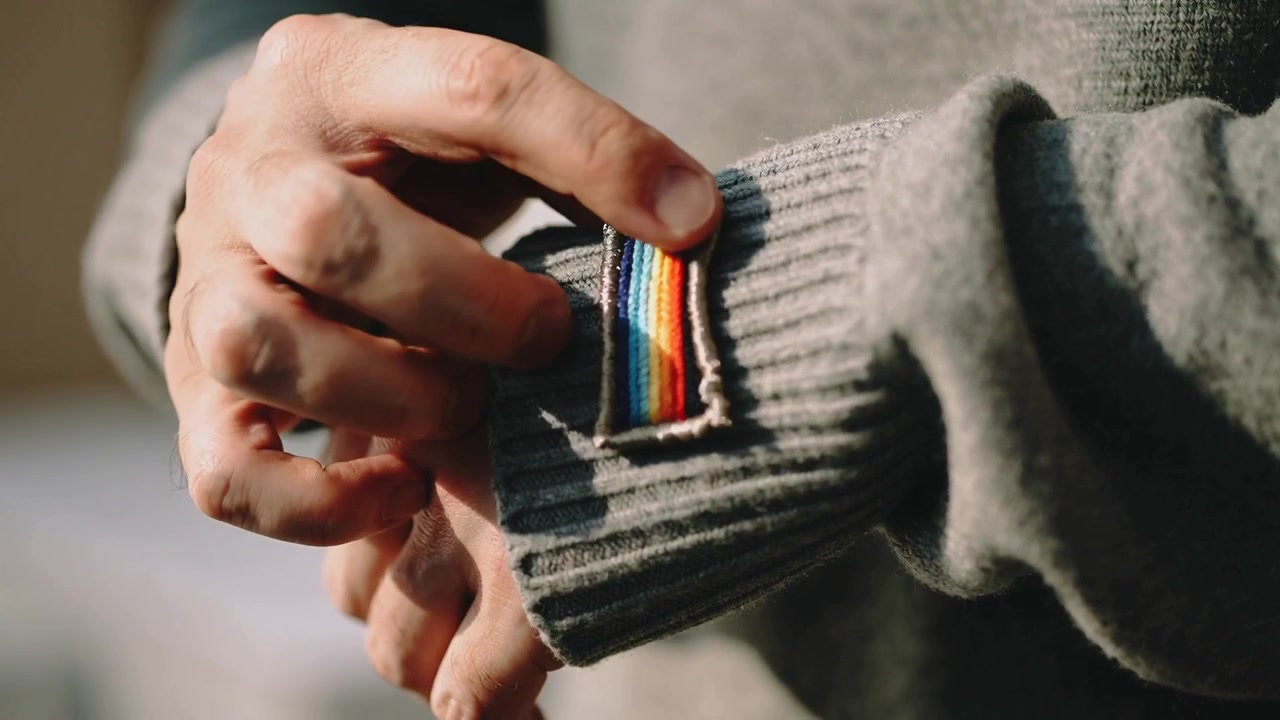 Mani di un uomo che liscia, il pollice destro che pizzica una toppa arcobaleno sul polsino di maglia grigia a casa per allineare i bordi.
