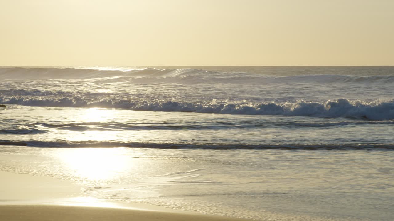 A panning shot follows waves rolling onto the beach, their foamy edges tracing patterns along the shore.