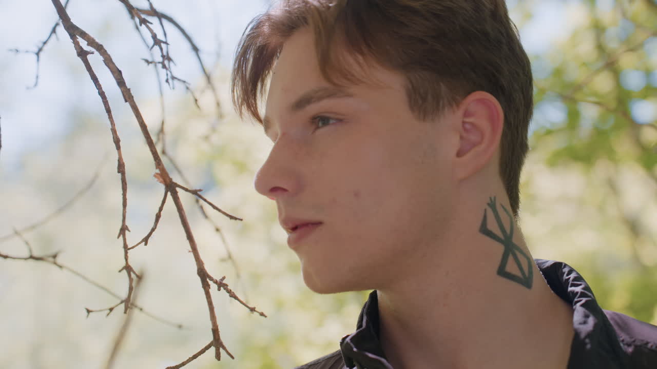 Young man in forest looks intently to right and back again, alert and observant with tattoo clearly visible on neck, sunlight filtering through trees, with serious expression and contemplation