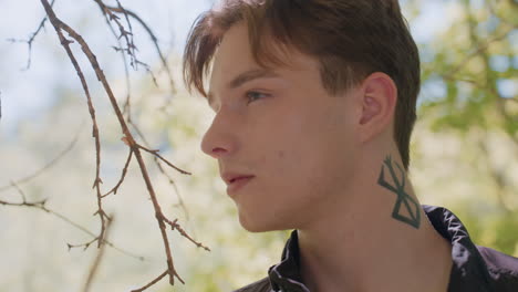 young man in forest looks intently to right and back again, alert and observant with tattoo clearly visible on neck, sunlight filtering through trees, with serious expression and contemplation